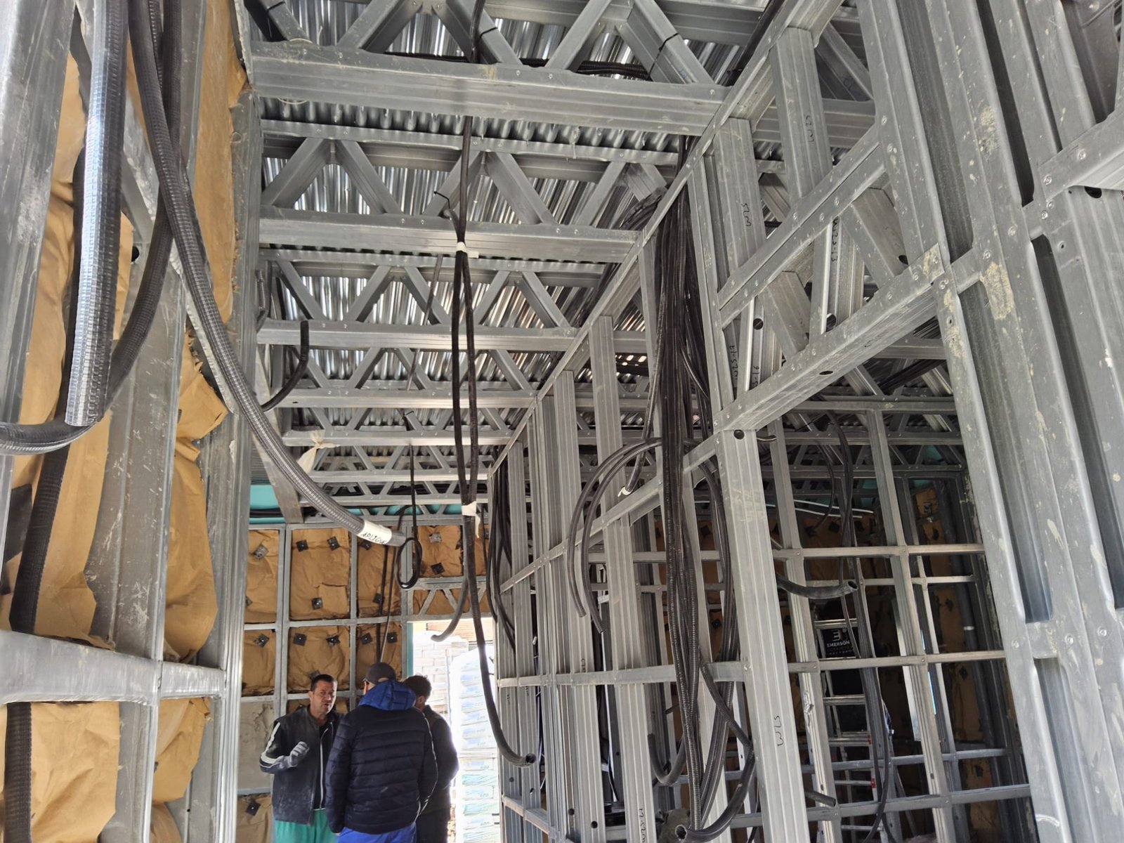 Vista interior de una construcción Steel Frame, mostrando la estructura metálica de vigas de celosía en el techo y montantes de pared. Numerosos cables eléctricos negros y conductos cuelgan y pasan a través de los perfiles de acero. Al fondo, se ven operarios y material aislante en las paredes.