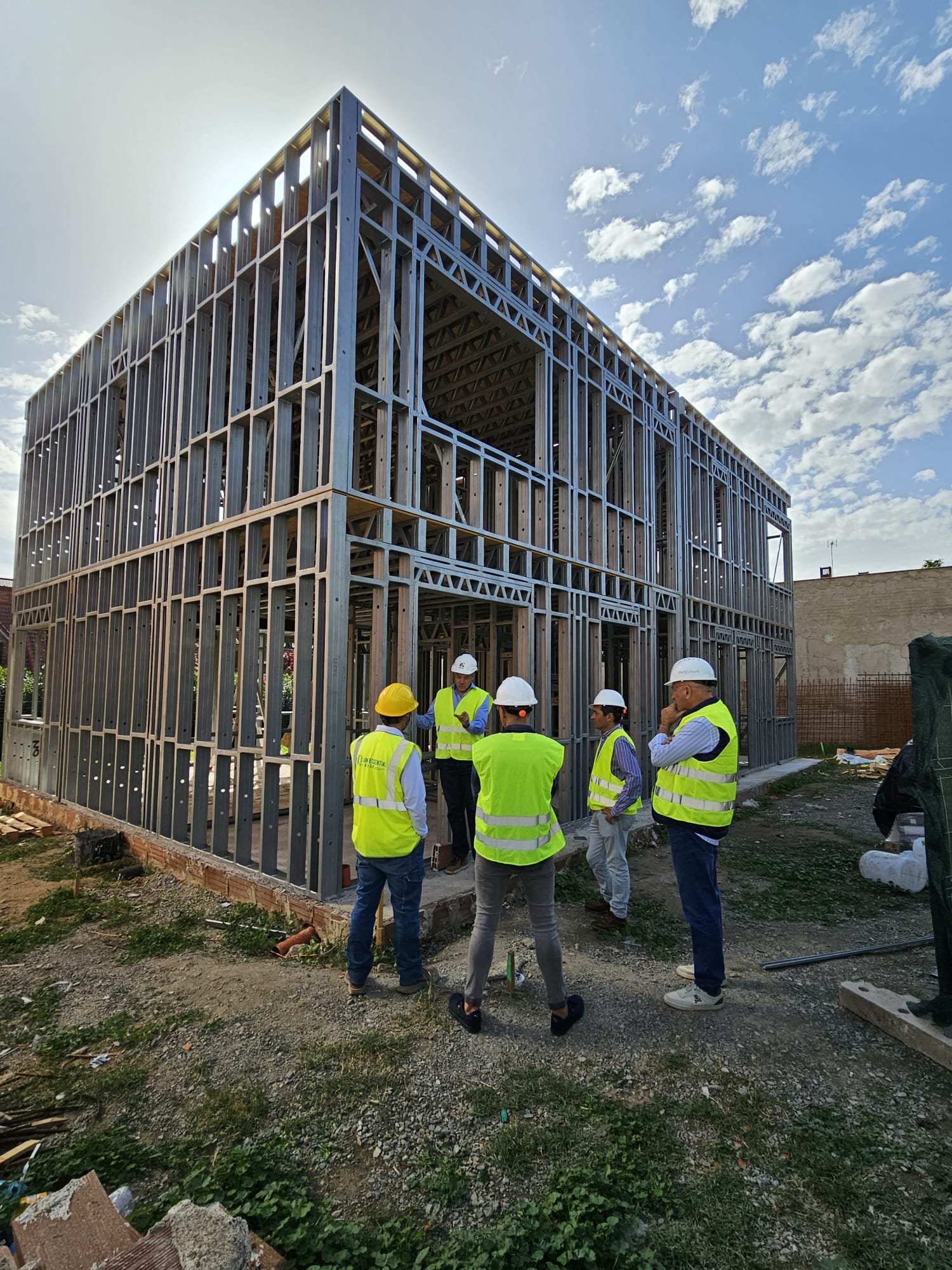 Un grupo de cinco ingenieros y responsables de obra, equipados con cascos y chalecos de alta visibilidad, mantienen una reunión de seguimiento frente a la estructura completa de acero ligero de una vivienda de dos plantas bajo un cielo despejado.
