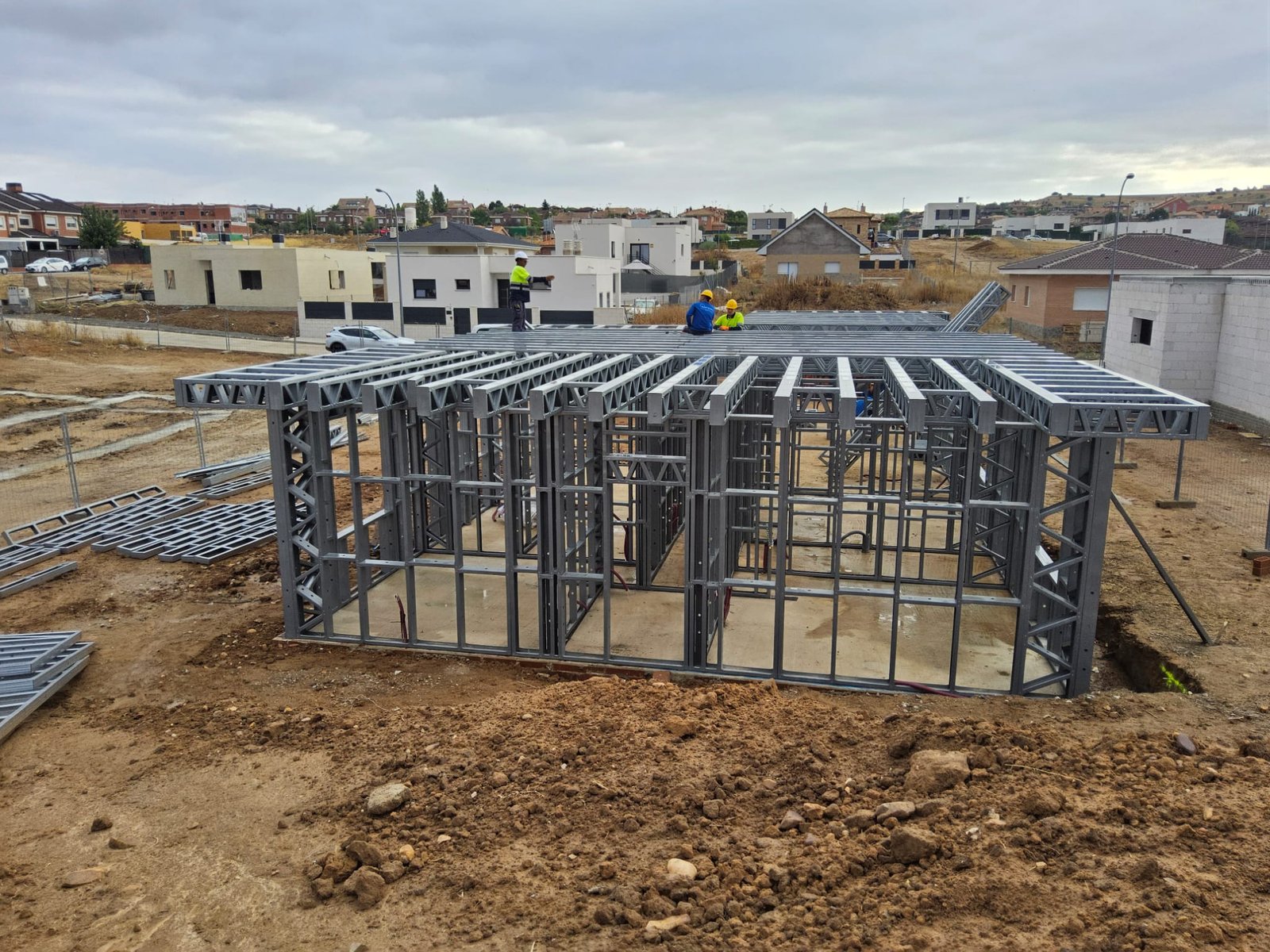 Vista de obra con la estructura de acero ligero de la planta baja completada. Dos operarios con chalecos reflectantes y cascos de seguridad trabajan en la instalación de las vigas del forjado superior sobre un terreno de tierra.